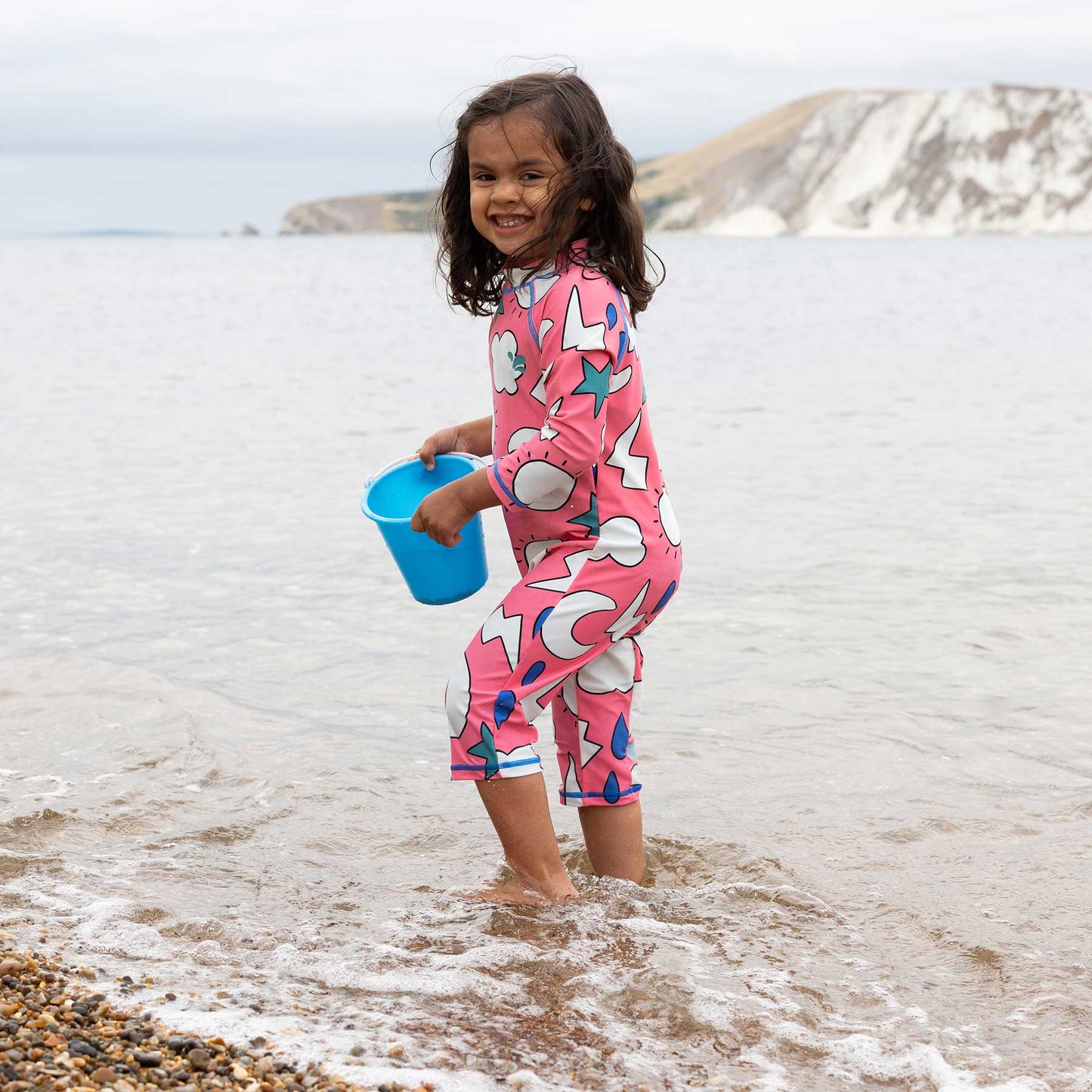 Girl wearing UV Protective Surf Suit Pink Weather, standing on a beach with a blue pail, enjoying the water in a sunsafe, durable swimwear.