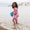 Girl wearing UV Protective Surf Suit Pink Weather, standing on a beach with a blue pail, enjoying the water in a sunsafe, durable swimwear.