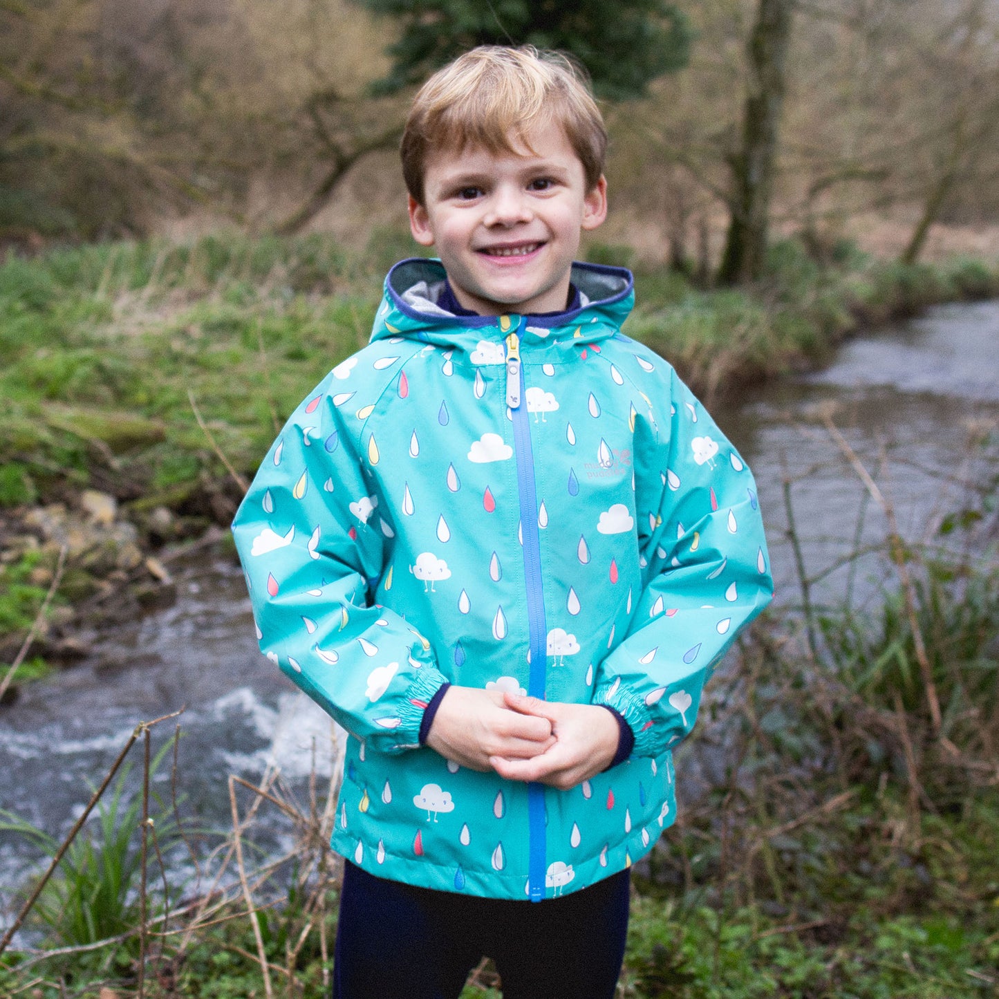 A young boy smiles, wearing an EcoLight Lightweight Waterproof Jacket Green Raindrops, stood near a stream in a leafy, natural setting.