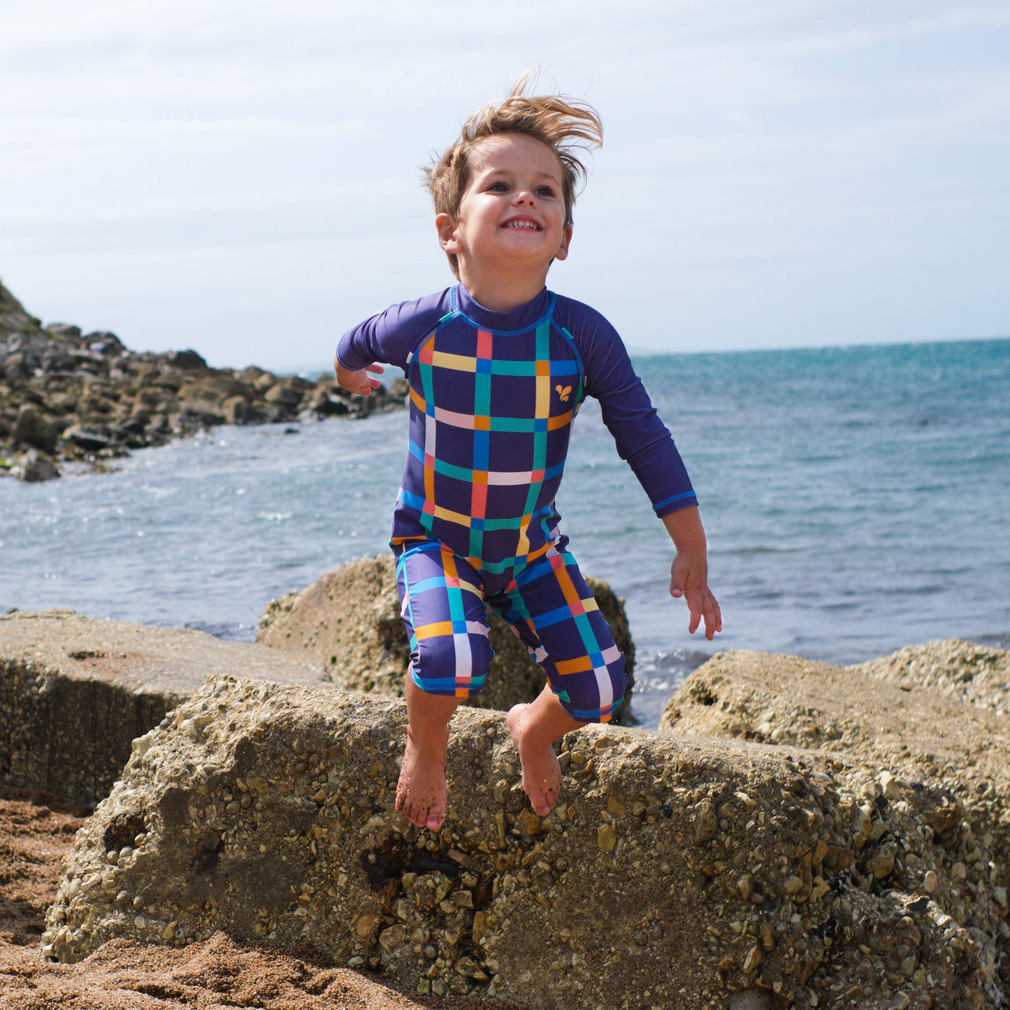 A young boy joyfully leaps off rocks at the beach, wearing a UV Protective Surf Suit Multi Check.