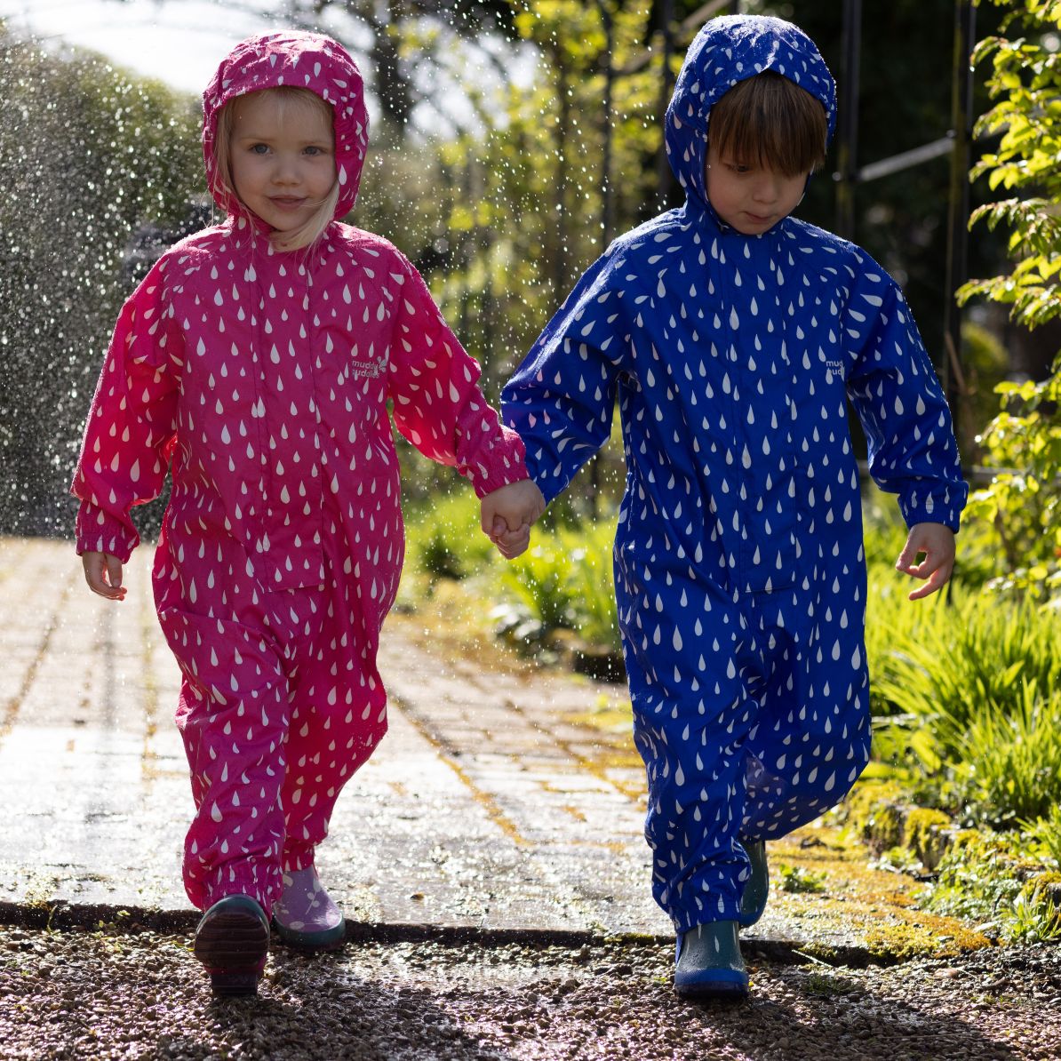 Two toddlers in Essentials Waterproof Puddle Suits Blue Raindrop explore a garden path, showcasing the durable, waterproof design ideal for rainy outdoor adventures.