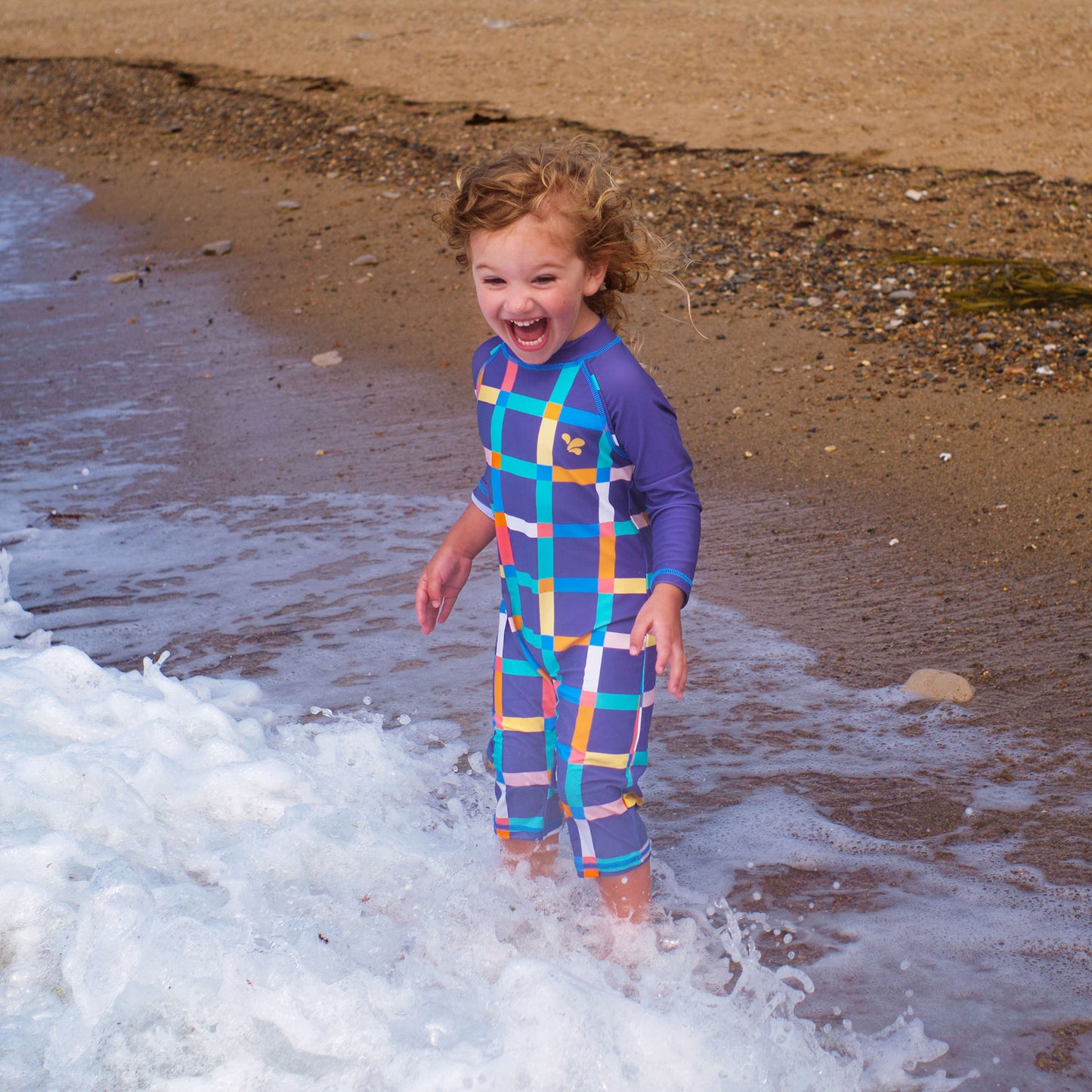 A toddler in a UV Protective Surf Suit Multi Check laughs while playing in the waves on a sandy beach.