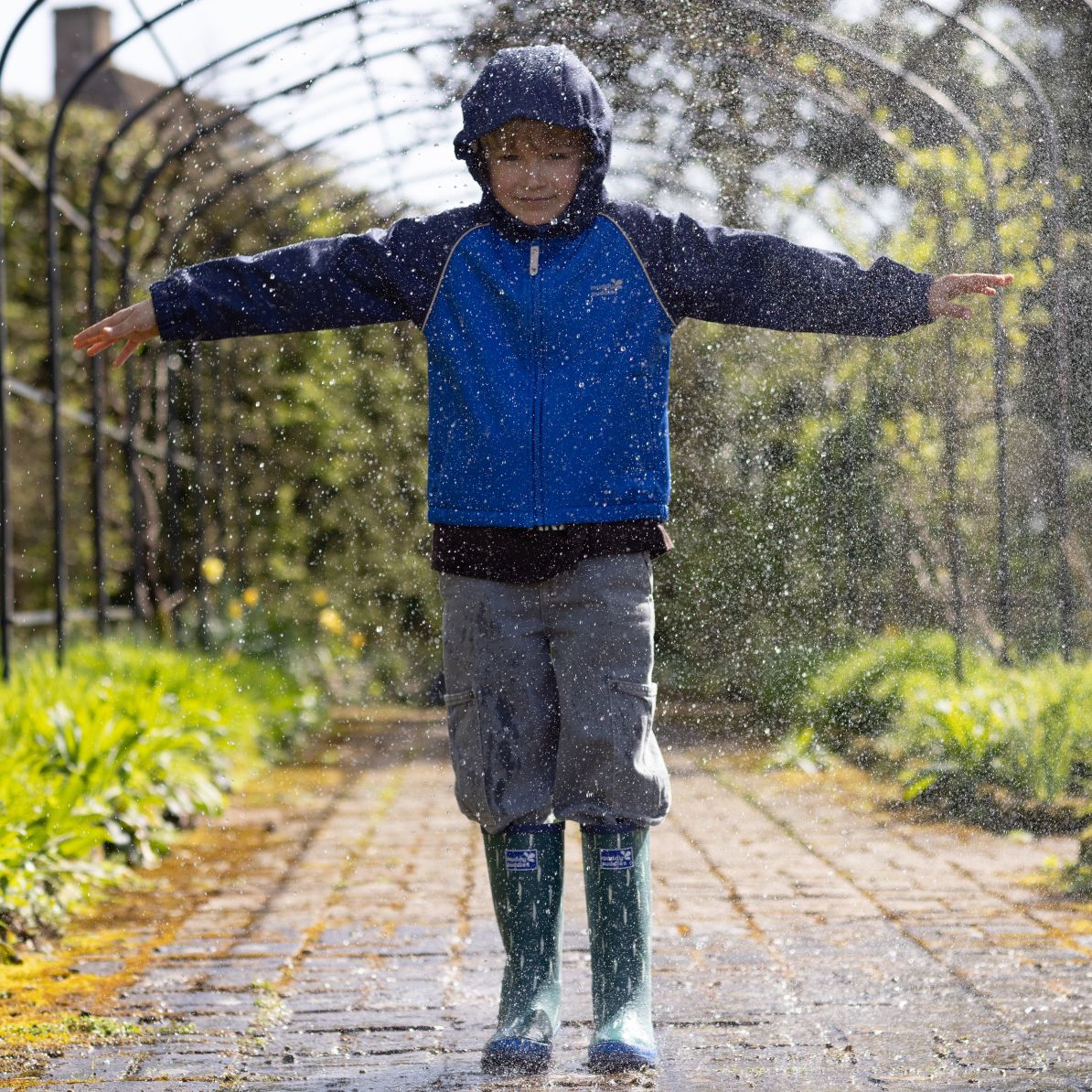 Child enjoying water spray in a durable Waterproof Softshell Jacket Blue, featuring elasticated cuffs, chin guard, and reflective details for outdoor adventures.