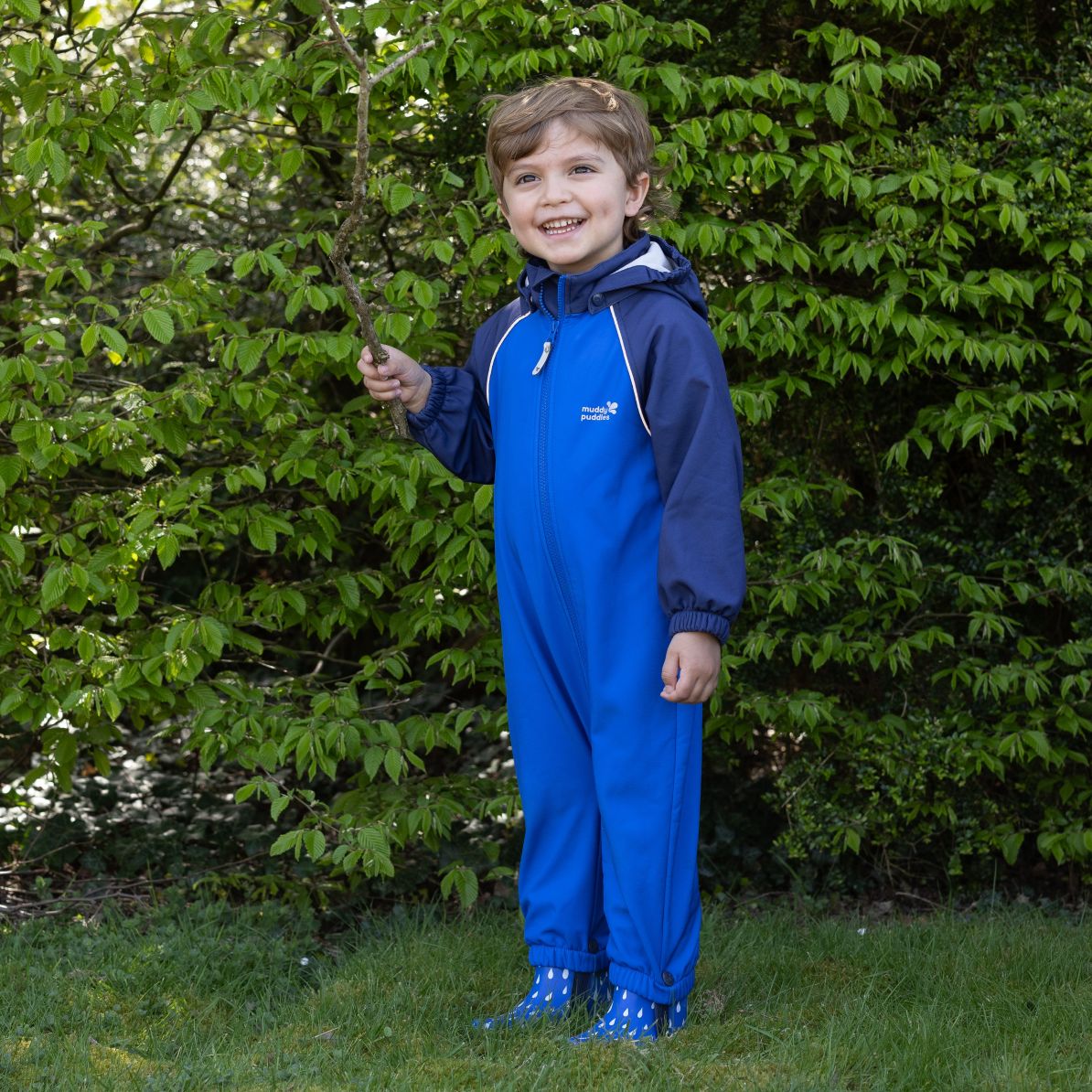 Child in a durable, waterproof blue softshell puddle suit holding a stick, smiling on grass, showcasing Muddy Puddles' high-performance children's outdoor wear.